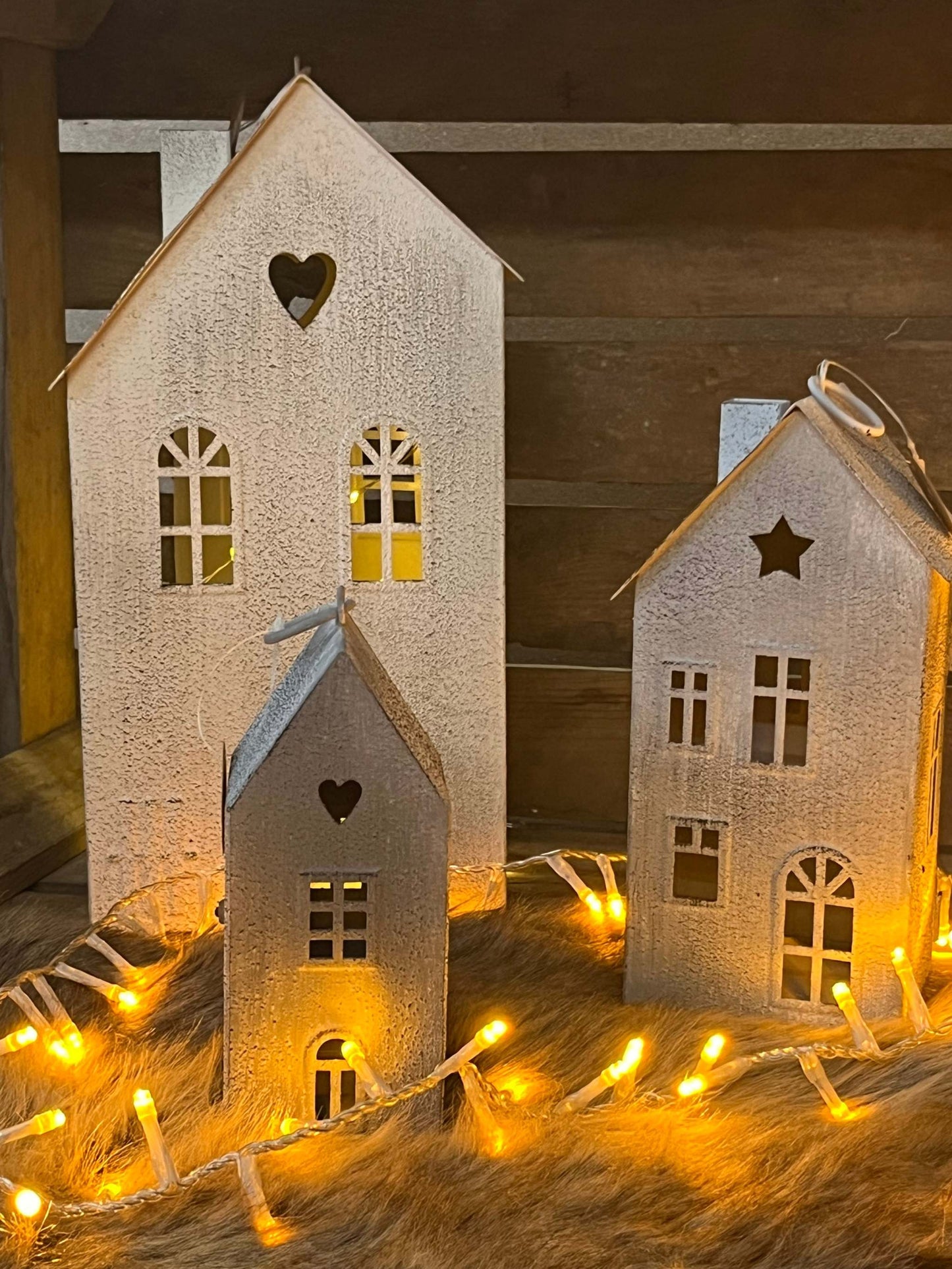 Metal miniature houses set with cut-out windows and heart-shaped decorations, lit by string lights in an antique white and gray finish.